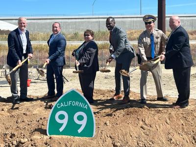 Six state and local officials pose for a photo while digging dirt with ceremonial shovels behind a green and white "California 9