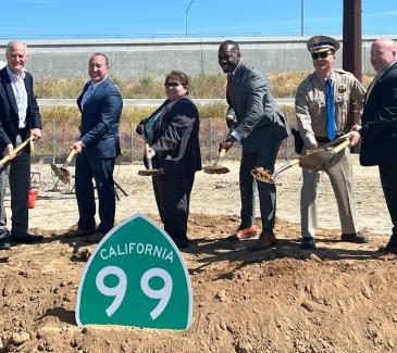 Six state and local officials pose for a photo while digging dirt with ceremonial shovels behind a green and white "California 9